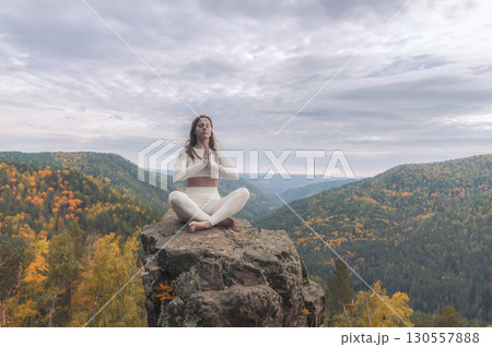 A white woman sits in the lotus position on a stone cliff, deep in meditation. Surrounded by breathtaking autumn landscapes, her serene presence harmonizes with the vibrant hues of the mountains. 130557888