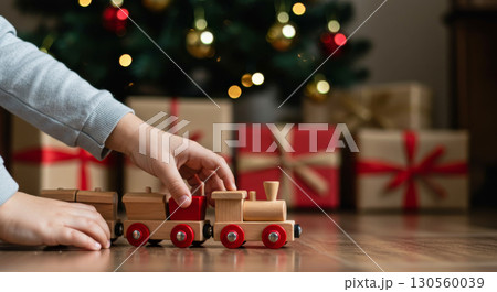 A child's hand plays with a wooden toy train on floor, with Christmas gifts and a festive tree in the background. 130560039