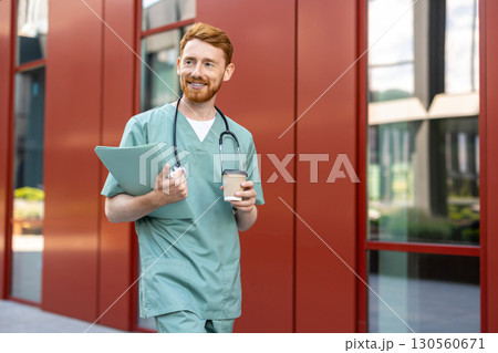 Man doctor with beard walking in scrubs holding coffee and folder Man doctor with beard walking in scrubs holding coffee and folder 130560671