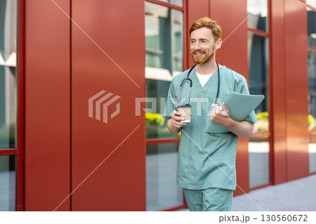 Male medic carrying coffee cup and documents near hospital building 130560672