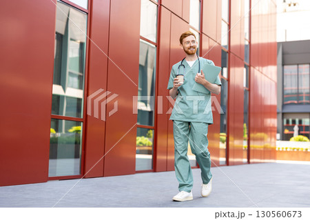 Man doctor in scrubs walking with takeaway coffee and paper folder Man doctor in scrubs walking with takeaway coffee and paper folder 130560673