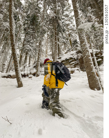 Person hiking through deep snow in winter forest wearing yellow jacket and backpack. 130561057