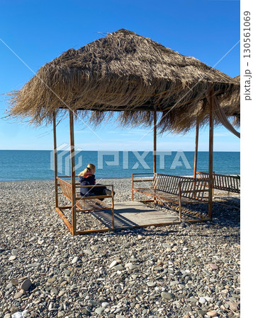 A woman sits alone under a rustic thatched gazebo on a pebble beach. In the background is a calm sea 130561069