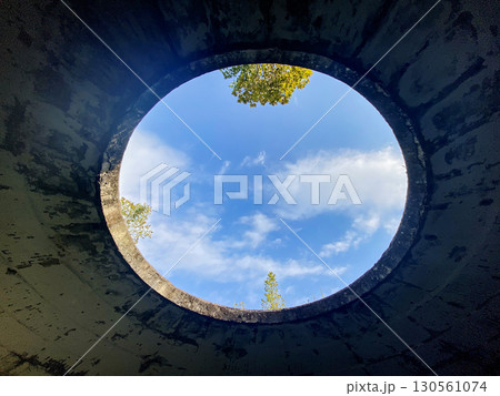 A round hole in a cracked concrete ceiling reveals a view of blue sky with clouds and treetops 130561074