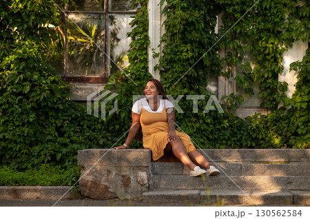Happy woman in mustard dress sitting on steps near greenery Happy woman in mustard dress sitting on steps near greenery 130562584