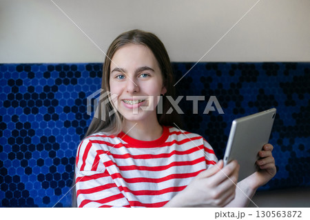 Teen girl wearing striped shirt smiles as she watches something on her tablet while seated in train Teen girl wearing striped shirt smiles as she watches something on her tablet while seated in train 130563872