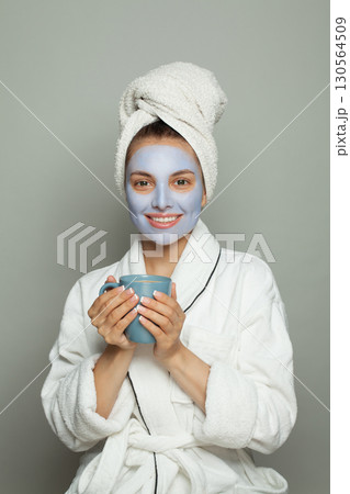 Smiling young woman with cosmetic mask on her face holding cup of morning coffee in hand Smiling young woman with cosmetic mask on her face holding cup of morning coffee in hand 130564509