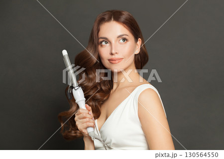 Young woman with long wavy hairstyle using hair curling tongs iron, indoor portrait. 130564550
