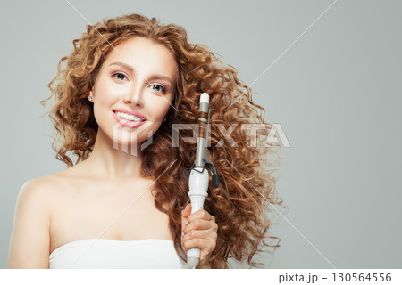 Happy woman with long wavy hairstyle using hair curling tongs iron, indoor portrait.  130564556
