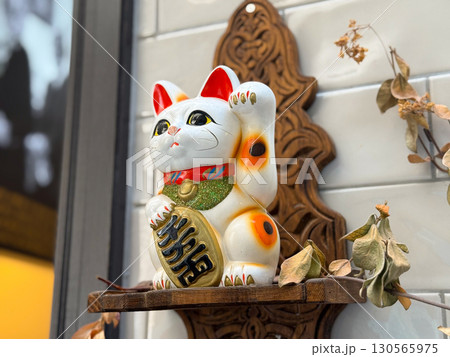 Maneki neko figurine with raised paw on wooden shelf. Tradition, prosperity, and cultural belief symbol in everyday lifestyle and decor. Maneki neko figurine with raised paw on wooden shelf. Tradition, prosperity, and cultural belief symbol in everyday lifestyle and decor. 130565975