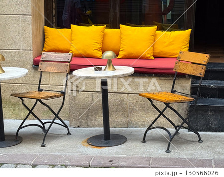 Outdoor cafe with wooden chairs, marble tables, and bright yellow pillows. Hospitality, comfort, and lifestyle in an urban social setting. Outdoor cafe with wooden chairs, marble tables, and bright yellow pillows. Hospitality, comfort, and lifestyle in an urban social setting. 130566026