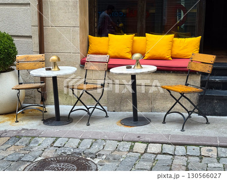 Outdoor cafe with wooden chairs, marble tables, and bright yellow pillows. Hospitality, comfort, and lifestyle in an urban social setting. 130566027
