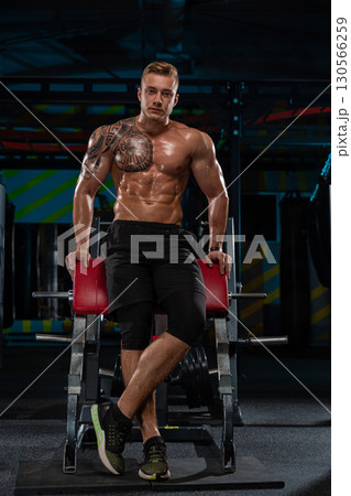 muscular man sits on a gym bench exuding confidence and strength. His toned physique and inked arm highlight his dedication to fitness. The background features gym equipment and vibrant lighting. muscular man sits on a gym bench exuding confidence and strength. His toned physique and inked arm highlight his dedication to fitness. The background features gym equipment and vibrant lighting. 130566259