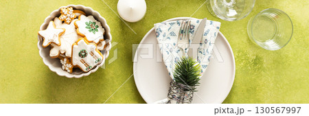 A festive Christmas table setting with an empty plate, cutlery, and napkin on a bright green background, decorated with spruse and white tree ornaments, banner 130567997