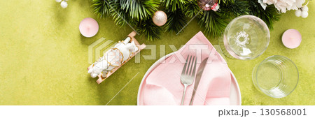 Top view of an elegant Christmas table with an empty plate, folded pink napkin and cutlery on a vibrant green background. Spruse branches and pink tree decorations add seasonal charm, banner 130568001