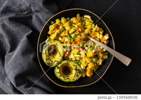 A colorful salad combines boiled egg, tender chicken breast, diced potatoes, vibrant carrots, and fresh green peas, topped with creamy avocado and sesame seeds, all drizzled with olive oil 130568036
