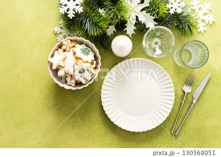 A bright green Christmas table setting with an empty plate and cutlery. The table is decorated with spruse branches and white tree ornaments, creating a festive vibe 130568051