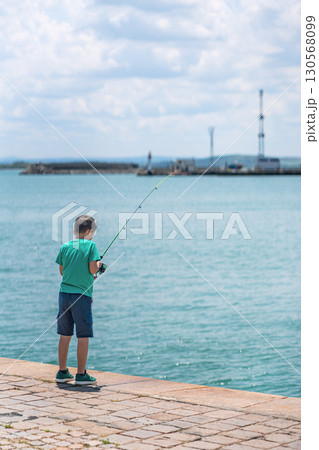 Young boy fishing alone by calm sea on sunny day, enjoying peaceful outdoor moment near harbor. Concept of childhood leisure, fishing activity, coastal relaxation Young boy fishing alone by calm sea on sunny day, enjoying peaceful outdoor moment near harbor. Concept of childhood leisure, fishing activity, coastal relaxation 130568099