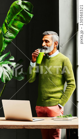 A man in a green sweater enjoys matcha while standing in a bright coworking space. Lush plants surround the area, creating a tranquil environment for productivity 130568321