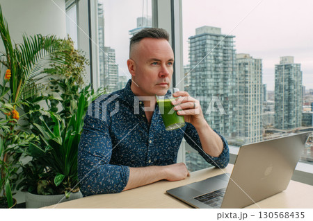 A man sips matcha from a green mug at a coworking space. He is focused on his laptop, with lush plants and sunlight creating a serene atmosphere 130568435