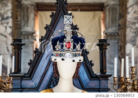 Mannequins in an ornate hall showcase embroidered gowns with pearls and feathered headpieces, reflecting regal ceremonial fashion. 130569953
