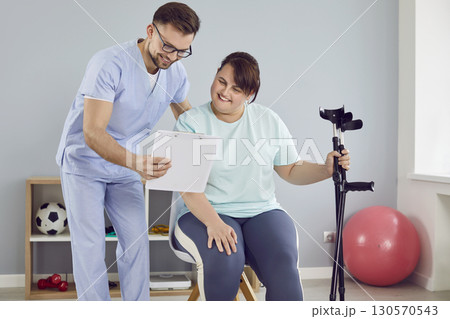Young nurse showing fat young woman with crutches a report file with appointment. Young nurse showing fat young woman with crutches a report file with appointment. 130570543