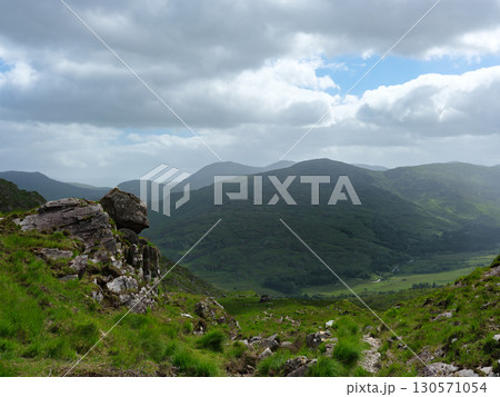 A vista across the Gap of Dunloe in Ireland shows lush green hills and valleys under a cloudy sky. Rock formations are visible on the hillside in the foreground. 130571054