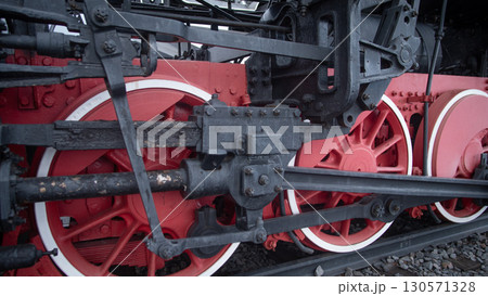 A close-up view highlights the colorful wheels and mechanical components of a vintage steam locomotive at a historical railway site during the day. The bright red contrasts with the dark metal. A close-up view highlights the colorful wheels and mechanical components of a vintage steam locomotive at a historical railway site during the day. The bright red contrasts with the dark metal. 130571328