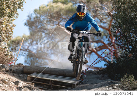 Mountain biker jumping high, wearing protective gear during intense enduro race, navigating wooden ramp with professional skill 130572543
