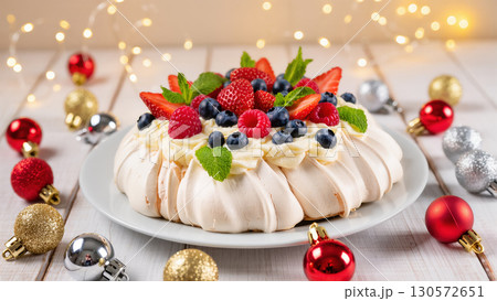 Christmas pavlova dessert topped with whipped cream, fresh berries, and mint leaves on a white plate, surrounded by festive ornaments and lights. Elegant holiday sweet. 130572651