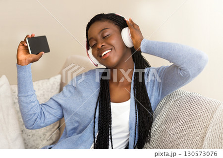 Favourite Song. Cheerful African American Girl In Headphones Listening Music On Smartphone And Singing On Sofa At Home Favourite Song. Cheerful African American Girl In Headphones Listening Music On Smartphone And Singing On Sofa At Home 130573076