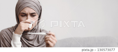 Unhappy arab woman blowing runny nose and looking at thermometer, having seasonal flu, closeup portrait with free space Unhappy arab woman blowing runny nose and looking at thermometer, having seasonal flu, closeup portrait with free space 130573077