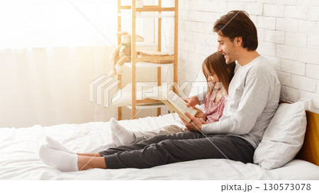 Father and little daughter spending weekend together at home, reading book, sitting on bed, copy space 130573078