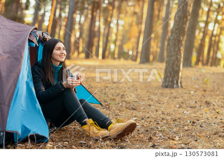 Peaceful young woman sitting in tent, drinking tea and looking around in forest, copy space 130573081
