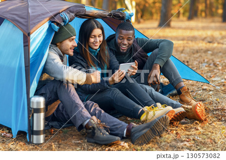Joyful international friends hikers resting at tent, watching photos on smartphone and drinking tea, copy space Joyful international friends hikers resting at tent, watching photos on smartphone and drinking tea, copy space 130573082
