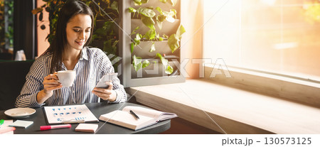 Working on business charts. Young millennial woman looking at smartphone and drinking coffee, making marketing analysis at cafe, free space 130573125