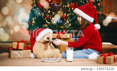 Lovely girl in Santa hat feeding Christmas teddy bear with chocolate cookies and milk on xmas eve 130573210