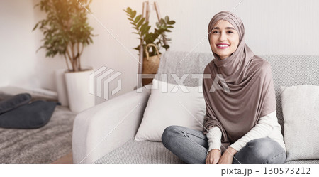 In Home Comfort. Beautiful Islamic Girl In Hijab Sitting On Couch And Smiling To Camera, Relaxing In Living Room, Resting On Sofa. 130573212