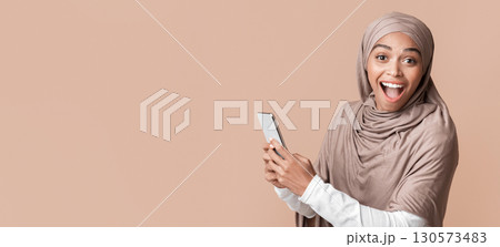 Unexpected message. Portrait of excited afro muslim girl in hijab holding smartphone and looking at camera, yellow background with free space 130573483