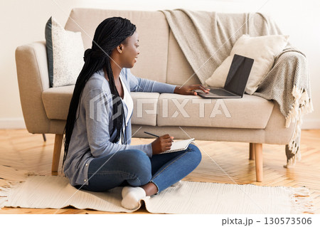 Young black woman watching webinar on laptop and taking notes while sitting on floor at home 130573506