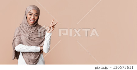 Cheerful black muslim girl in hijab showing peace gesture or v-sign and looking at camera, posing over yellow studio background, free space 130573611