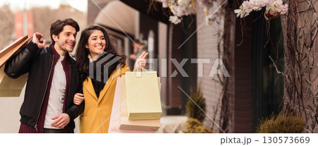 A couple smiles at each other while holding shopping bags and a credit card, walking along a bustling city street in autumn, copy space A couple smiles at each other while holding shopping bags and a credit card, walking along a bustling city street in autumn, copy space 130573669