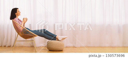 Time To Relax. Smiling girl drinking hot tea, sitting in wicker chair and looking out of window at home, free space 130573696