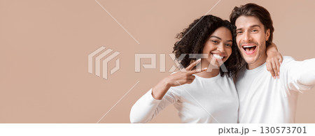 Cheerful millennials taking selfie. Joyful interracial couple capturing self-portrait, posing together over yellow background, girl gesturing peace 130573701