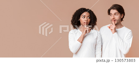 Common secret. Interracial couple holding fingers on lips, making shh sign, hiding some information, yellow studio background with empty space 130573803
