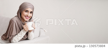 Portrait of happy muslim girl in hijab relaxing at home with cup of coffee, sitting on sofa and looking at camera, copy space Portrait of happy muslim girl in hijab relaxing at home with cup of coffee, sitting on sofa and looking at camera, copy space 130573804