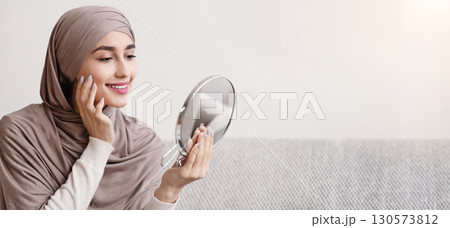 Beauty routine. Attractive muslim girl in hijab holding mirror and looking at her face with perfect soft skin, sitting on sofa at home, free space Beauty routine. Attractive muslim girl in hijab holding mirror and looking at her face with perfect soft skin, sitting on sofa at home, free space 130573812