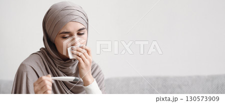 Cold And Flu. Sick muslim woman in hijab sitting on sofa looking at thermometer, and blowing her nose, feeling unwell at home, closeup Cold And Flu. Sick muslim woman in hijab sitting on sofa looking at thermometer, and blowing her nose, feeling unwell at home, closeup 130573909