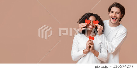 Falling In Love. Cheerful Man Holding Red Paper Hearts Over Eyes Of His Afro Girlfriend And Smiling At Camera, Yellow Background With Empty Space Falling In Love. Cheerful Man Holding Red Paper Hearts Over Eyes Of His Afro Girlfriend And Smiling At Camera, Yellow Background With Empty Space 130574277
