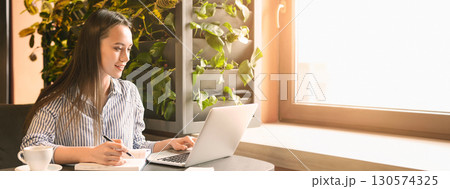 A woman sits at a table in a cafe, focused on her laptop while taking notes, surrounded by greenery and natural light, copy space A woman sits at a table in a cafe, focused on her laptop while taking notes, surrounded by greenery and natural light, copy space 130574325